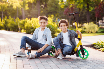 Active children's entertainment in the summer park. Two cute brothers having fun in the park on a summer evening. Riding on a skateboard and scooter. Dressed in denim suits and white T-shirts