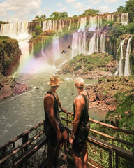 Gay travel couple in front of a paradisiac waterfall with rainbows and macaw