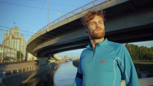 Lockdown medium shot of handsome young athlete finishing running workout, looking away thoughtfully and catching breath standing on bridge over river, headphones in his ears