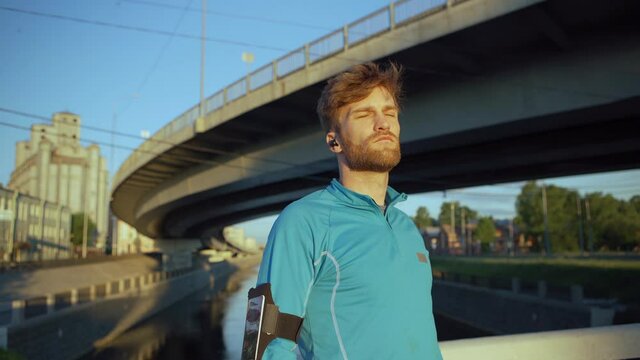 Lockdown medium shot of handsome young man finishing jogging cardio workout, looking away, catching breath and smiling standing on river over bridge. Headphones in his ears, cellphone armband on arm
