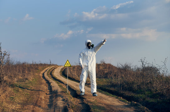 Environmentalist standing on the road near yellow triangle with skull and crossbones warning sign and pointing finger at sky. Research scientist wearing protective suit, gas mask and shoe covers.