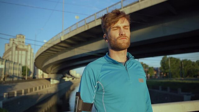 Lockdown medium shot of handsome young athlete looking away thoughtfully, breathing deeply and closing eyes standing on bridge over river and recovering after jogging workout