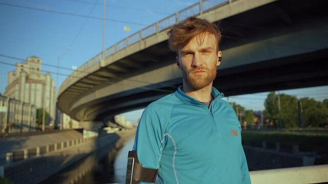 Medium shot portrait of handsome young athlete turning head and looking at camera standing on bridge over river and recovering after jogging workout
