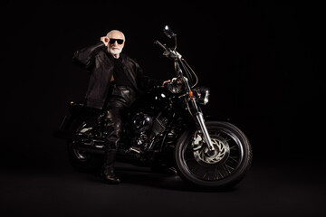 Fototapeta na wymiar Full size photo of old man confident cool biker man sit bike touch sunglasses specs wear modern outfit isolated over black color background