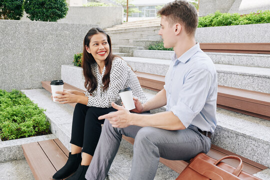 Smiling pretty young mixed-race woman looking at coworker telling intersting story when they are sitting outdoors