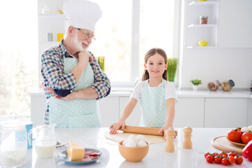 Obraz na płótnie Canvas Portrait of nice cheerful cheery glad grey-haired granddad teaching grandchild learning cooking fresh delicious dish kneading dough recipe ingredient cuisine modern light white interior kitchen house