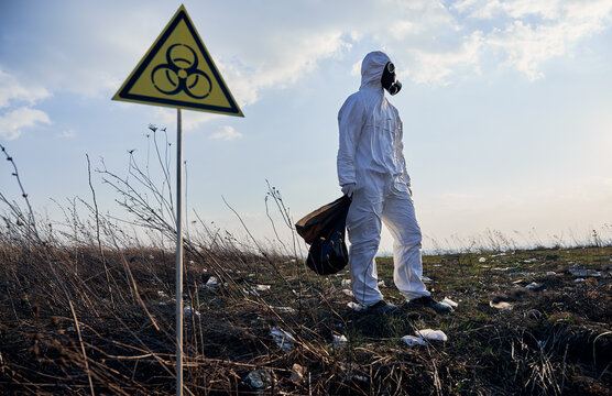 Full length of male ecologist in radiation suit and gas mask holding garbage bag, standing near biohazard warning sign on abandoned territory with trash. Concept of ecology, environmental pollution