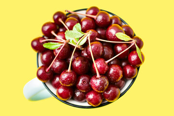 Close-up of fresh red ripe cherry with sprigs and leaves in a rustic mug, top view on yellow background. Sweet summer fruits and water droplets in a cup. Healthy seasonal food. Summertime vitamins.