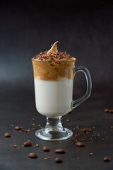 Glass of trendy fluffy creamy dalgona or whipped instant coffee with milk, coffee beans and chocolate on the dark background. New popular food and drink trend concept. Copy space.