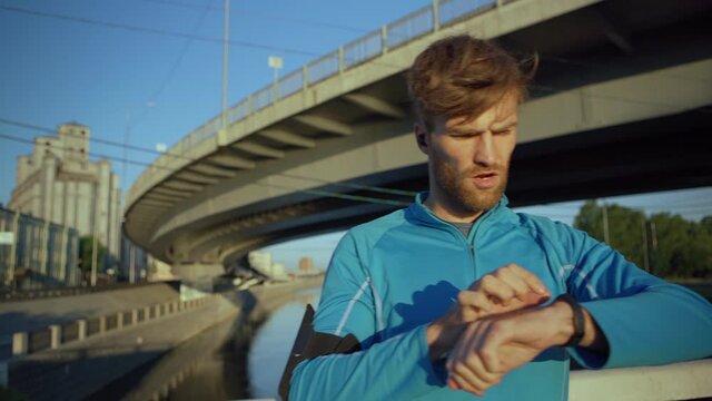Lockdown medium shot of tired young male athlete finishing jogging workout, checking time on smartwatch and catching breath standing on bridge over river, headphones in his ears
