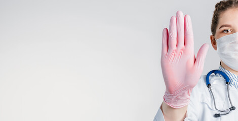 A young beautiful woman doctor in a white coat and pink sterile gloves shows a stop gesture on...