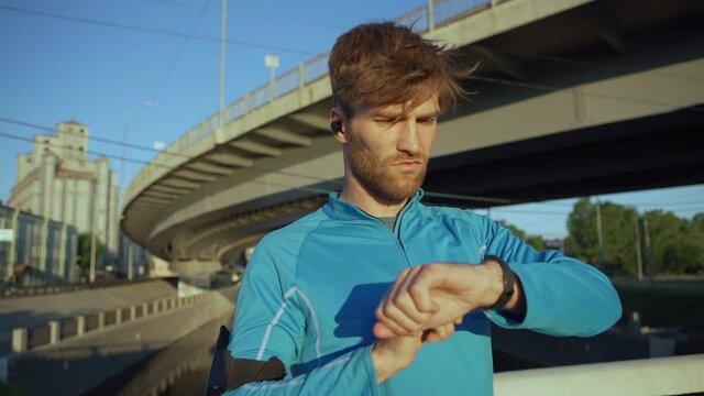 Waist up portrait of handsome male athlete looking around, checking time on smartwatch and smiling in slow motion while standing on bridge over river after jogging