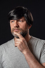 Portrait of attractive man in his 40s with grey and black beard and long black hairs. Black background. Male dressed in grey t-shirt. Looking away from the camera hand at his face.