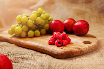 Still life, fresh juicy raspberries on a wooden board in focus. Green grapes and red nectarines out of focus, hessian backdrop.