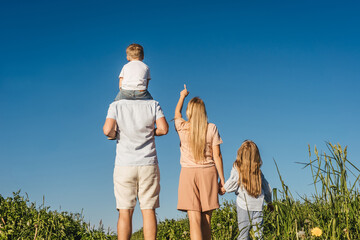 Happy family of mother, father and two children have a rest in field. Mother shows a finger to the sky and they looks up. Back view.