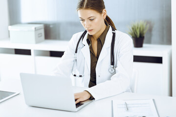 Woman-doctor typing on laptop computer while sitting at the desk in hospital office. Data in medicine and healthcare