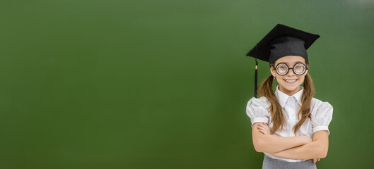 Nerd girl wearing graduation hat stands near empty chalkboard with crossed hands. Empty space for text