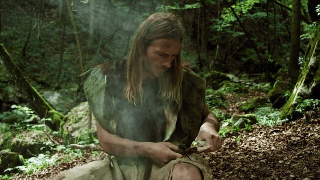 Prehistoric Man Sits In Forest Sharpening Wooden Spear With Stone
