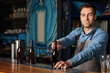 Serious handsome barman in apron opens bottle of beer in interior of modern pub