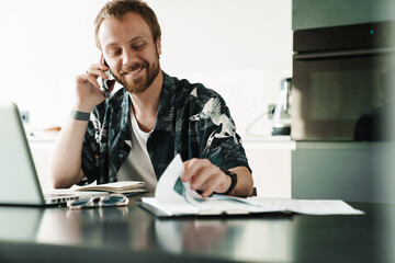 Photo of smiling man talking on cellphone while working with laptop