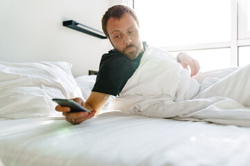 Photo of young bearded man using mobile phone while lying after sleep