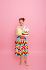 Red-haired girl in colorful dress on a pink background with sweet marshmallow cake. Vertical orientation. Woman holding cake in hands