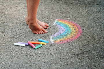 Children's feet on the asphalt next to a rainbow drawn with colored crayons