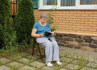 An elderly woman is sitting in the courtyard on a summer day and reading a book. The concept of a happy old age and International Day of Older Persons
