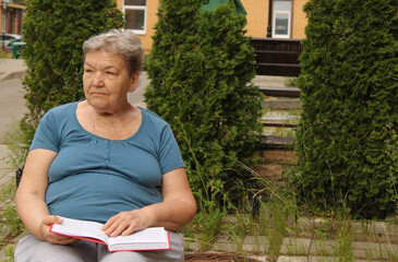 An elderly woman is sitting in the courtyard on a summer day, reading a book and sometimes thinking about something. The concept of a happy old age and International Day of Older Persons