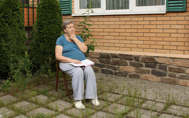 An elderly woman is sitting in the courtyard on a summer day, reading a book and sometimes thinking about something. Lifestyle. The concept of a happy old age and International Day of Older Persons