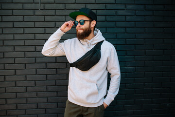 Handsome hipster guy wearing gray blank hoodie, black cap and black waist bag with space for your...
