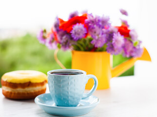 Cup of coffee, donut and beautiful bouquet of bright wildflowers in yellow watering can on white wooden background.