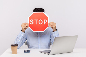 Man employee sitting in office workplace with laptop, hiding face behind stop symbol, warning with red traffic sign, symbol of prohibition restriction. indoor studio shot isolated on white background