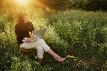 New office concept. Fashionable elegant girl working on laptop and sitting on rustic chair in warm sunset light in summer field. Remote work with social distancing and safety protocols