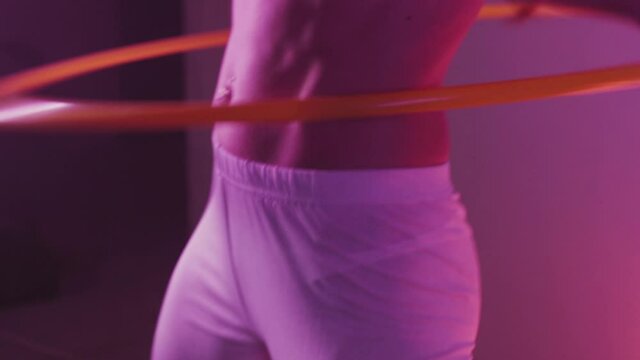 Athletic girl in white leggings twists a hoop on a muscular waist in violet light