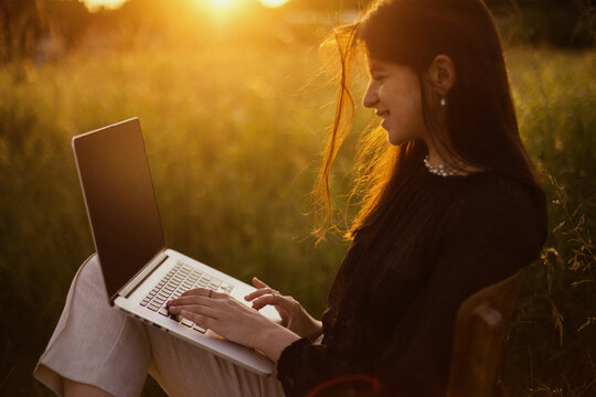 Freelance and remote work outdoors. Young business woman working online. Fashionable elegant girl with laptop sitting on rustic chair in sunny summer field at sunset. Creative image - Powered by Adobe