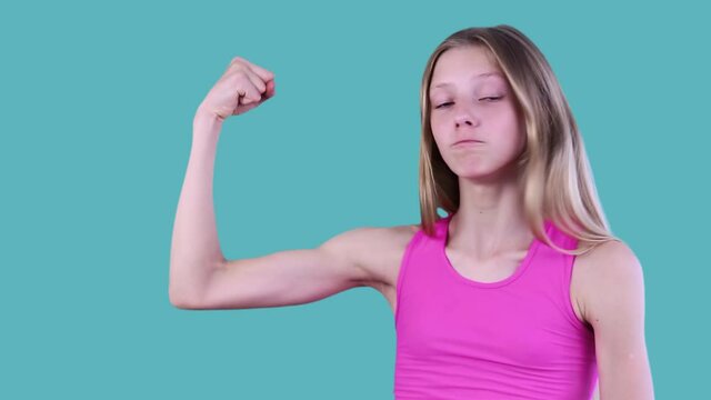 Beautiful Stylish Blond Girl In Pink T-shirt Showing Arm Muscles Symbolizes Strength, Feeling Joy And Pleasure, Demonstrating Thumb-up. Isolated On Blue Background. 