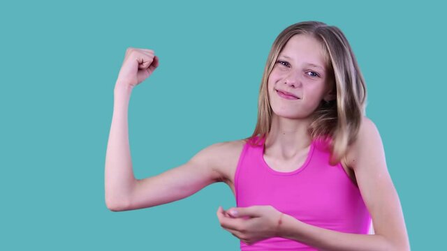 Close up portrait of an attractive thin teen girl with long blond hair demonstrates the lack of arm muscles, showing thumb down with disappointment over blue background. Healthy lifestyle concept.