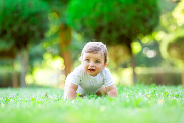 Naklejka premium Cute kid in white bodysuit learns to crawl. Healthy toddler crawls on green grass in summer