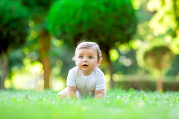 Cute kid in white bodysuit learns to crawl. Healthy toddler crawls on green grass in summer