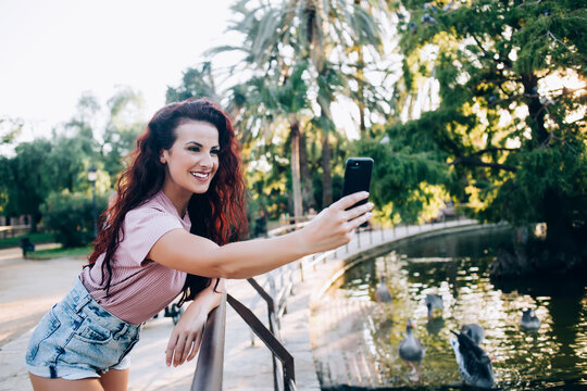 Beautiful brunette curly female influencer posing for photo on mobile phone resting in city park with lake on weekends,charming female 20s using smartphone for making selfie on free time vacations