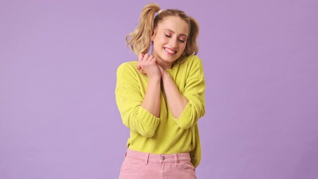A beautiful positive blonde modestly crosses her arms near her face, rejoicing at something in an isolated studio on a purple background