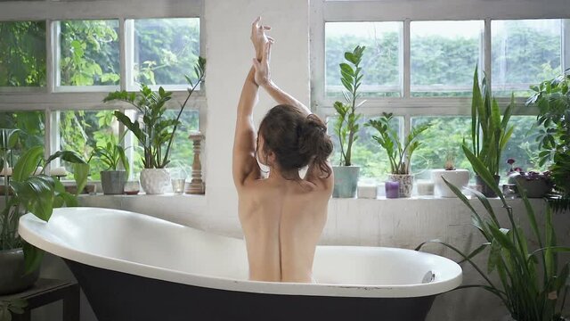 Brunette woman with naked back sitting in the bathtub, posing, touching her hands. Slowmotion. 