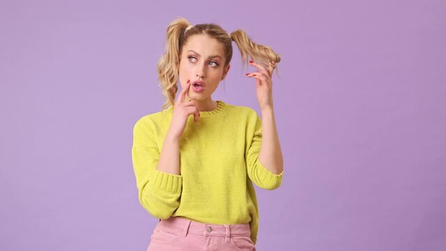 An unrivaled blonde girl thoughtfully puts hair on her finger, shows the index finger of her hand near her cheek in an isolated studio on a purple background