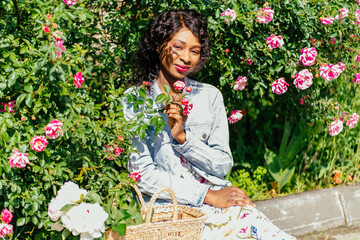 Outdoor portrait of thouughtful sensual black woman with in jeans jacket, elegant summer dress, straw bag with peonies posing in summer park near roses bushes. Summer mood concept.