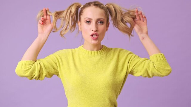 A cute blonde in a yellow sweater smiles at the camera and makes grimaces in an isolated studio on a purple background