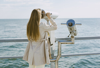 Young woman in beige trench coat stands by stationary binoculars and holds it with both hands. She stands with her back to the frame, looks towards the sea.
