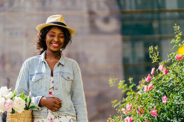 Black woman smiling in a flowering bush of red roses.Millennial black woman in wicker hat holding straw bag with peonies near pink rose bush, wear jeans jacket walking at city street. Summer mood conc