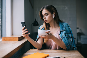 Stylish female student using smartphone and wireless earbuds while drinking tea in creative...