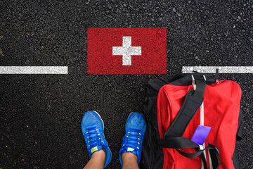 a man with a shoes and travel bag is standing on asphalt next to flag of Switzerland and border